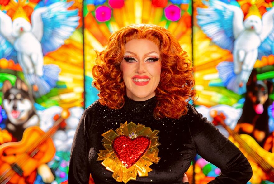 Cabaret performer Geraldine Quinn in costume and full make-up, stylised red hair, wearing a black top with a red padded heart stitched onto the front of it, posing  against a stained glass background.