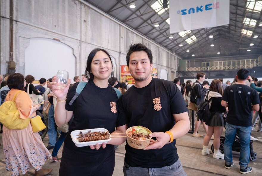 Two people at the Sake Festival, each holding take-away containers of food and one of them holding a glass in her hand; in the background people are walking around the Sake Festival inside a large hall.