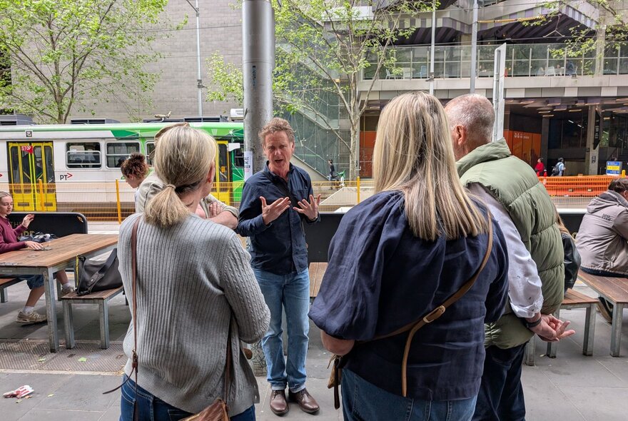 Small tour group standing beside outdoor tables, listening to a man speaking.