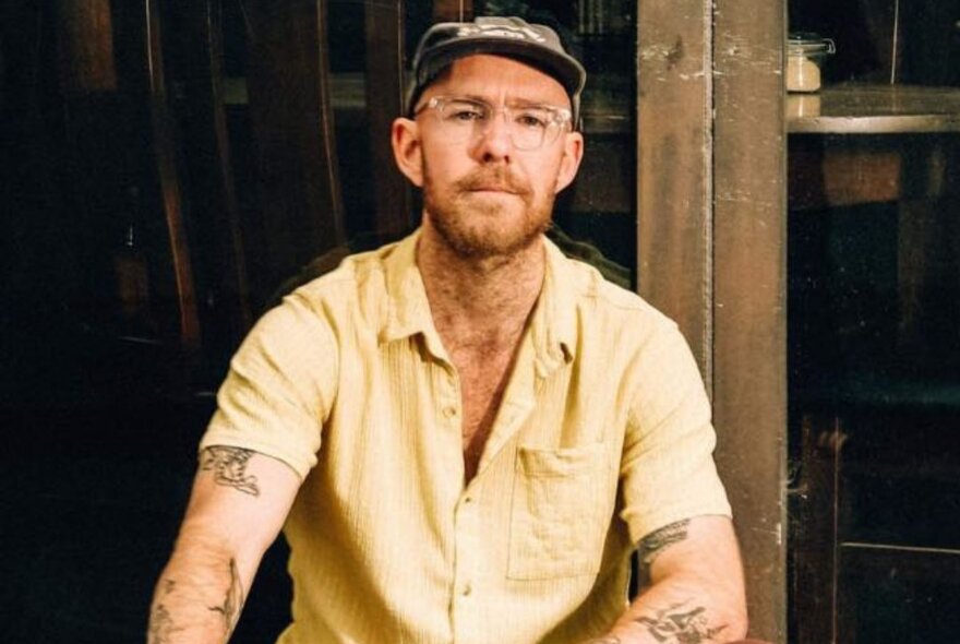 A man with a cap and tattoos in a casual shirt looking neutral. 