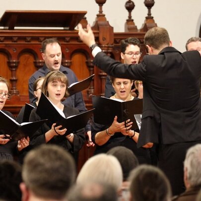 A choral performance, featuring a conductor leading a vocal ensemble, with a seated audience in the foreground. 
