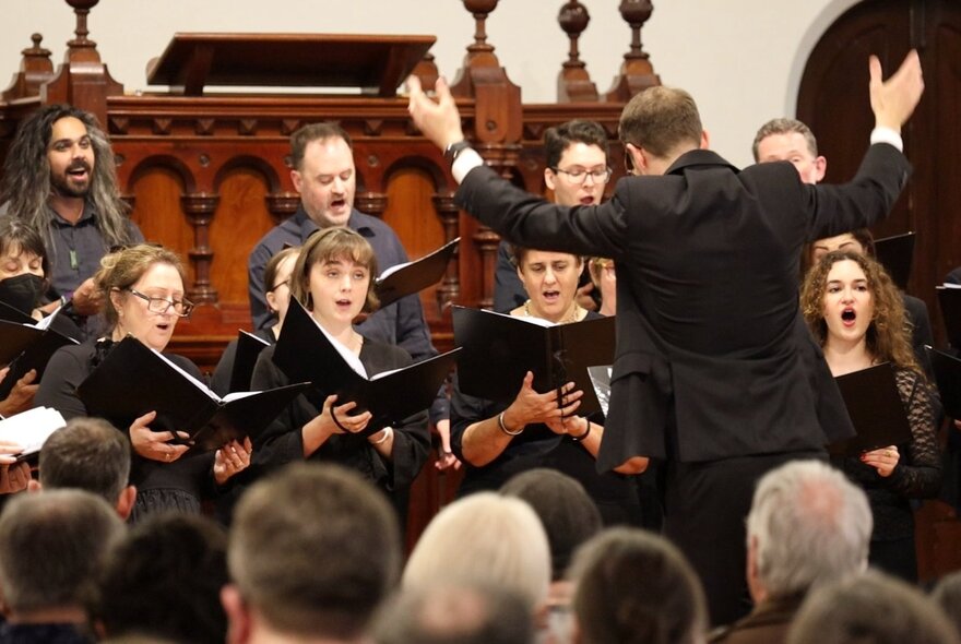 A choral performance, featuring a conductor leading a vocal ensemble, with a seated audience in the foreground. 