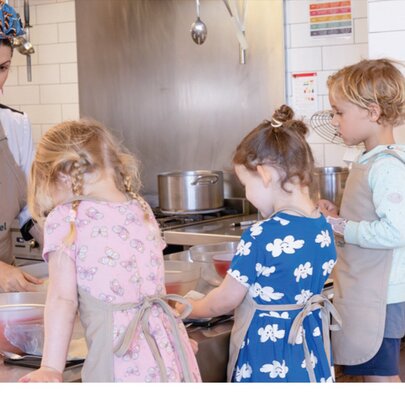 Children at a counter wearing aprons all learning to make pretzels with a woman wearing a headscarf.
