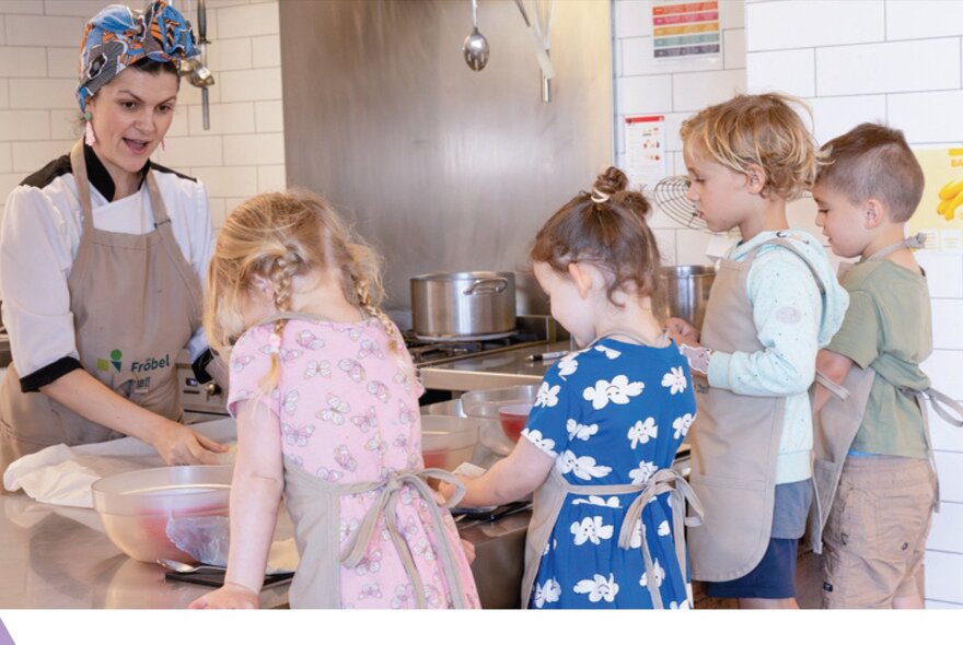Children at a counter wearing aprons all learning to make pretzels with a woman wearing a headscarf.