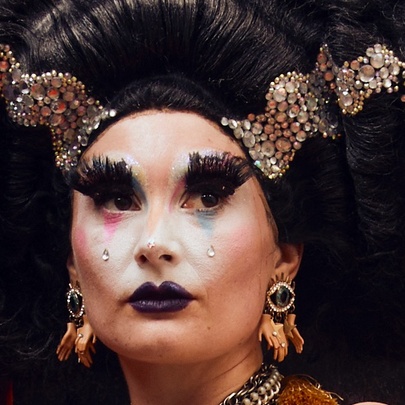 A person wearing heavy stage makeup with very exaggerated bushy eyelashes and a large black wig with ornate ribbons. 