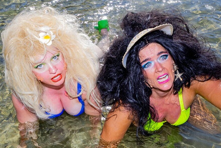 The two performers from the show BIRDS, both wearing bikinis and with big teased hair lying in the shallow water at the beach.