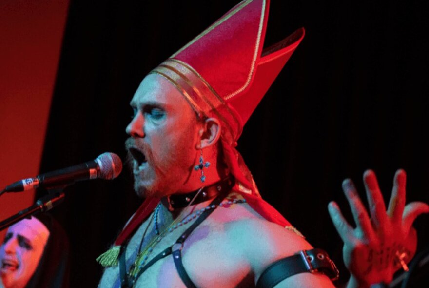 A performer on stage singing into a microphone and wearing a red bishop's tall hat, parts of a leather harness visible over his chest.