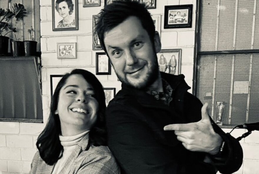 Man and woman standing back to back in a room with many pictures on the walls, the man pointing a finger at the woman looking up at him.