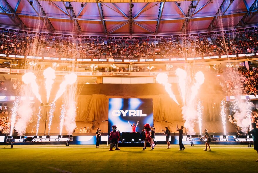 Pre-game entertainment at a BBL cricket match with jets of flames and a DJ on a small stage, with cricket fans in the stadium seats and the roof closed at Marvel Stadium.