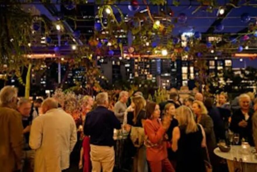 A crowd of people partying and socialising at Loop Rooftop garden, at night with city buildings in the background.