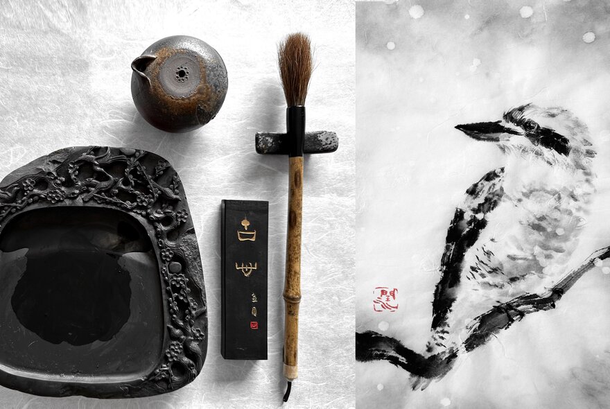An ink drawing of a kookaburra on a twig with a fine brush and a bowl of ink next to it.