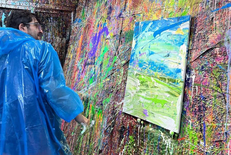 A  person in protective plastic clothing splashing paint onto a canvas on a paint splattered wall.