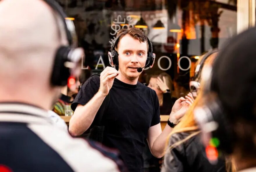A man in black with a microphone headset on, performing a walking comedy show to an audience with headphones.