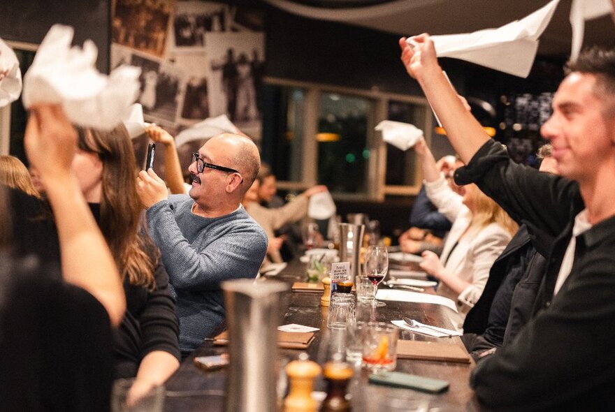 Seated dining patrons in a restaurant clapping and waving white napkins in the air above them, enjoying a musical performance. 