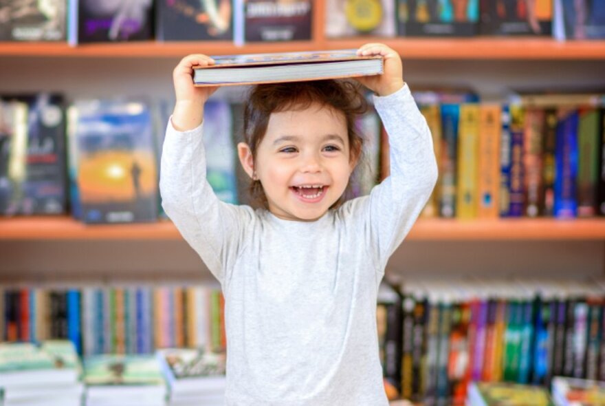 A smiling preschool aged child balancing a book on their head that they hold with both hands, in  a library setting.