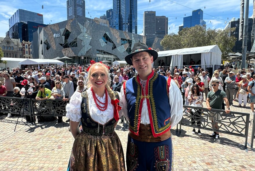 A couple wearing Polish national costume, with a crowd of people behind them at the Polish Festival in the open plaza at Fed Square on a bright sunny day.  