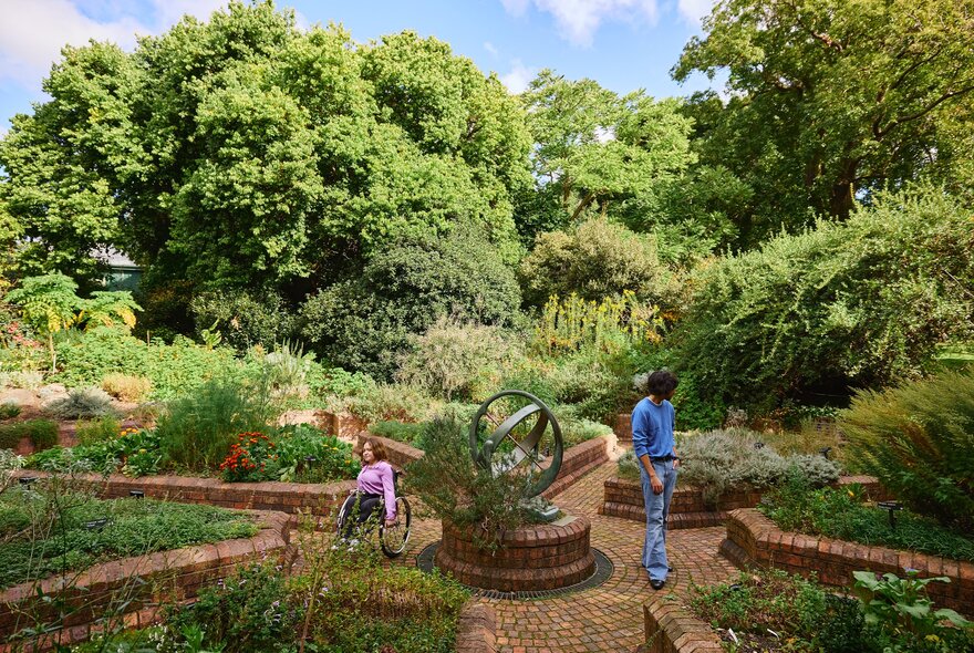 Two friends are in a paved garden with garden beds filled with shrubs. One friend is standing the other is in a wheelchair. 