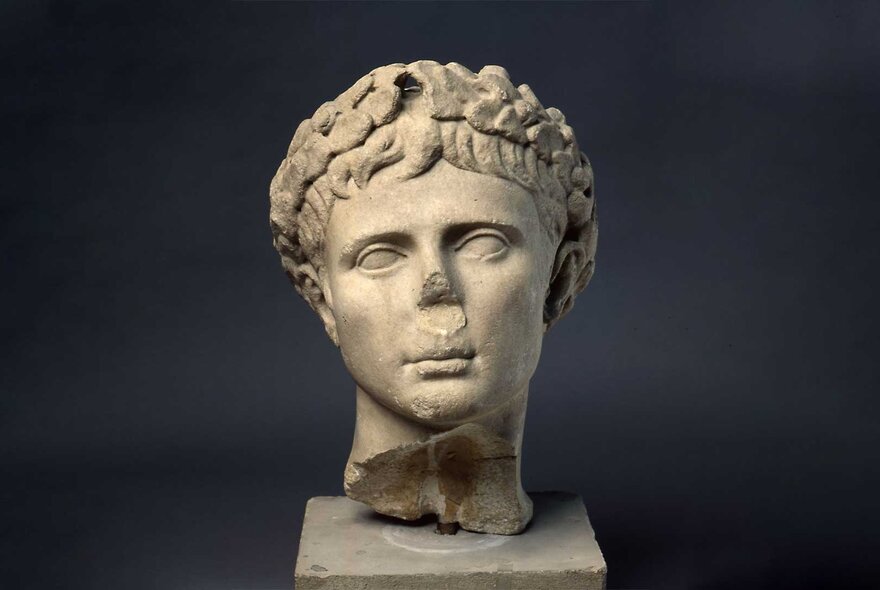 An ancient Roman marble bust with its nose chipped off, displayed on a small square plinth.