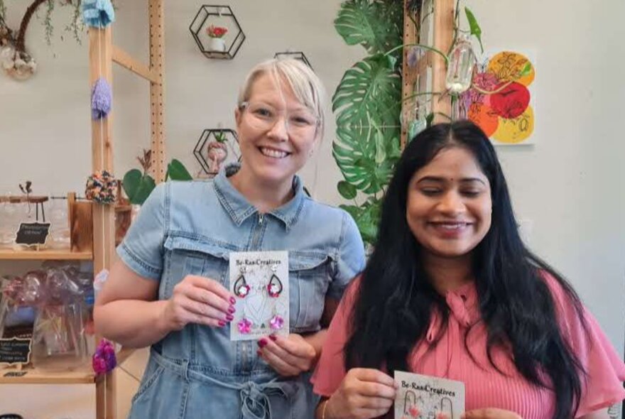 Two smiling workshop participants standing in a studio, holding up jewellery they have made.