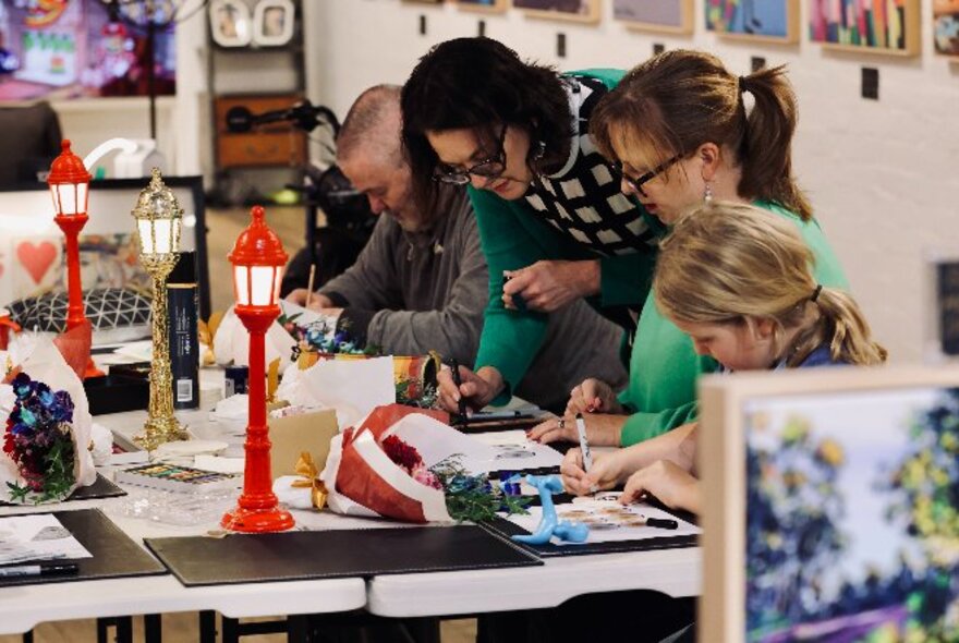 People at a table interacting and painting in an art workshop, while their tutor leans over behind them and offers  instruction.