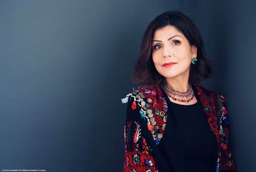 Author Samah Sabawi, an elegantly dressed woman with shoulder-length straight dark hair, wearing a beaded necklace, posed against a dark grey backdrop.