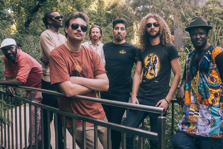 A group of seven musicians posing on a small balcony or bridge in a garden, looking serious.