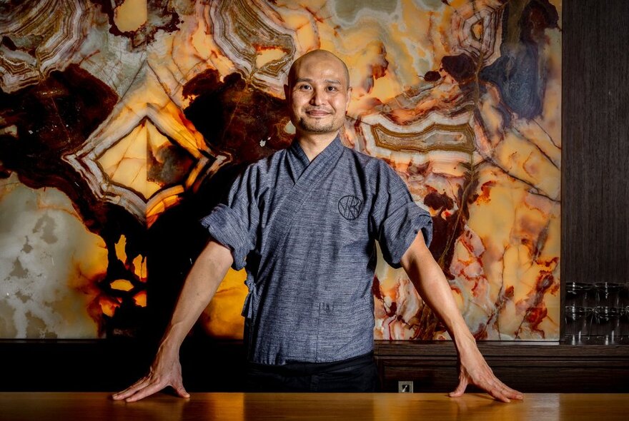 A sushi chef looking pleased and leaning his hands on a table with artwork in the background. 