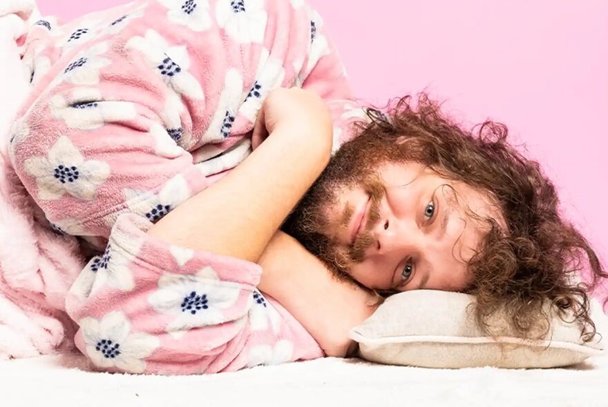A person lying down in a pink and white floral patterned dressing gown, resting on a pillow with a pink background.