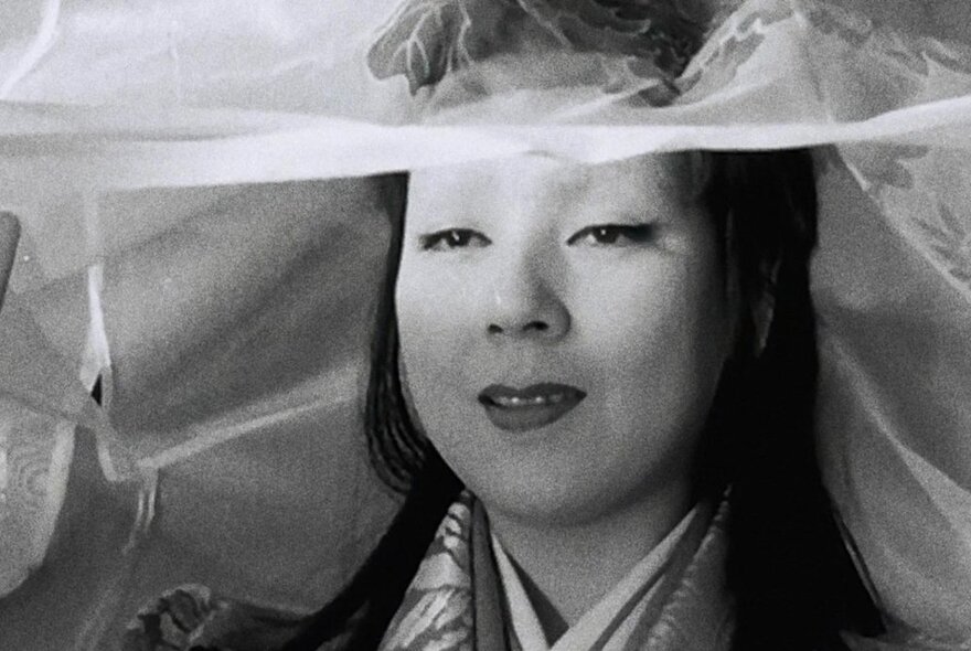 Black and white still from a Japanese horror film of a geisha's pale face, smiling serenely, a thin veil resting on her head.