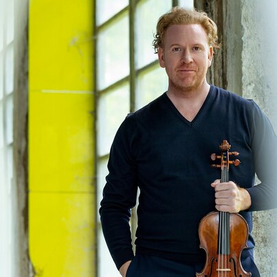 Violin virtuoso Daniel Hope, posing with his violin in front of a wall of glass bricks and a yellow column.