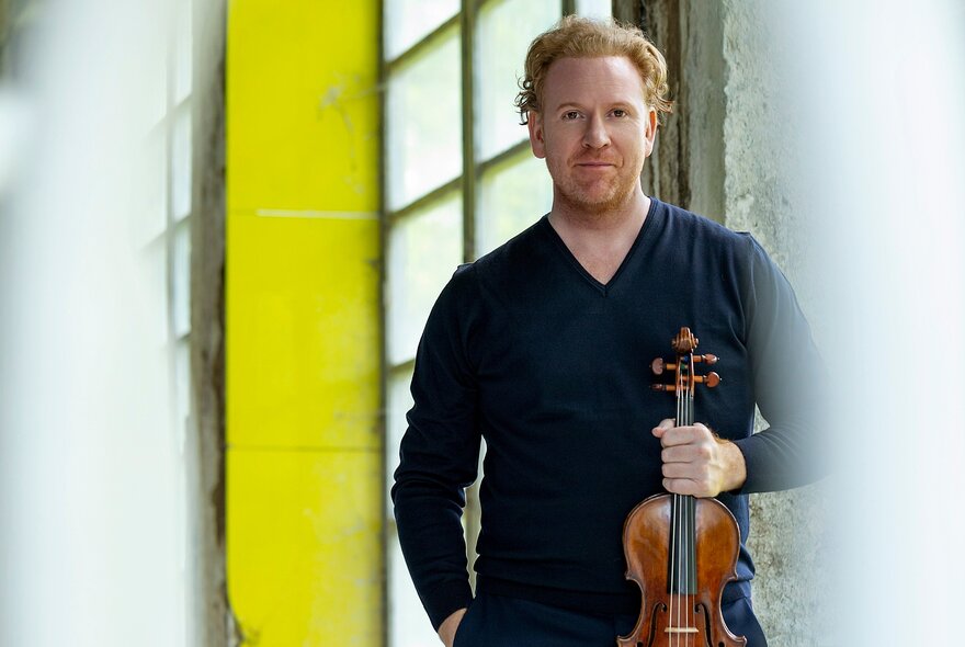 Violin virtuoso Daniel Hope, posing with his violin in front of a wall of glass bricks and a yellow column.