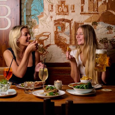 Two women enjoying a meal at Hofbräuhaus with plates of food and glasses of wine and beer laid out before them.