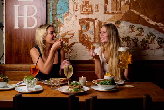 Two women enjoying a meal at Hofbräuhaus with plates of food and glasses of wine and beer laid out before them.