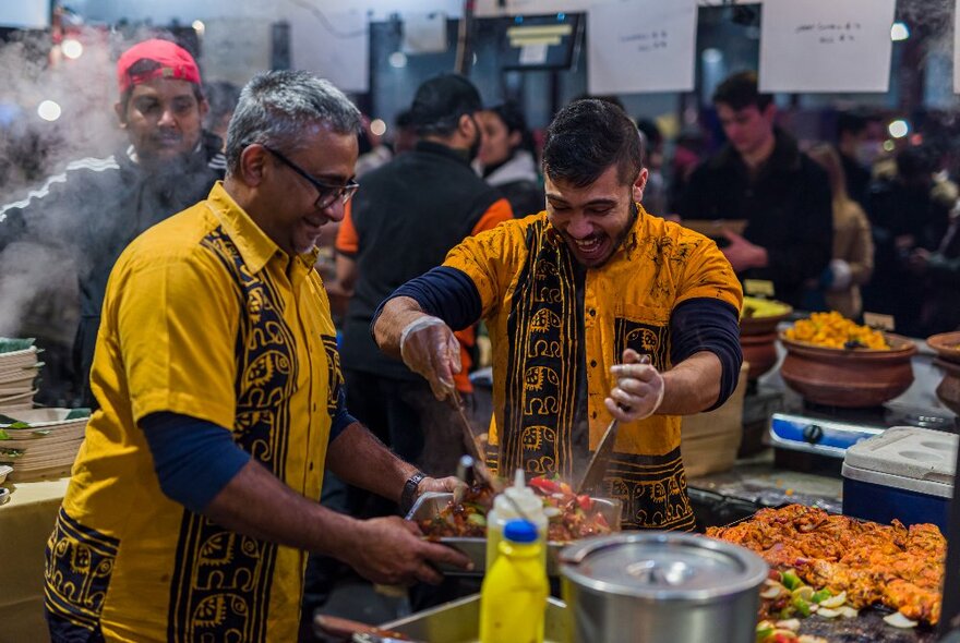 Men in yellow shirts cooking at a market with people looking on. 
