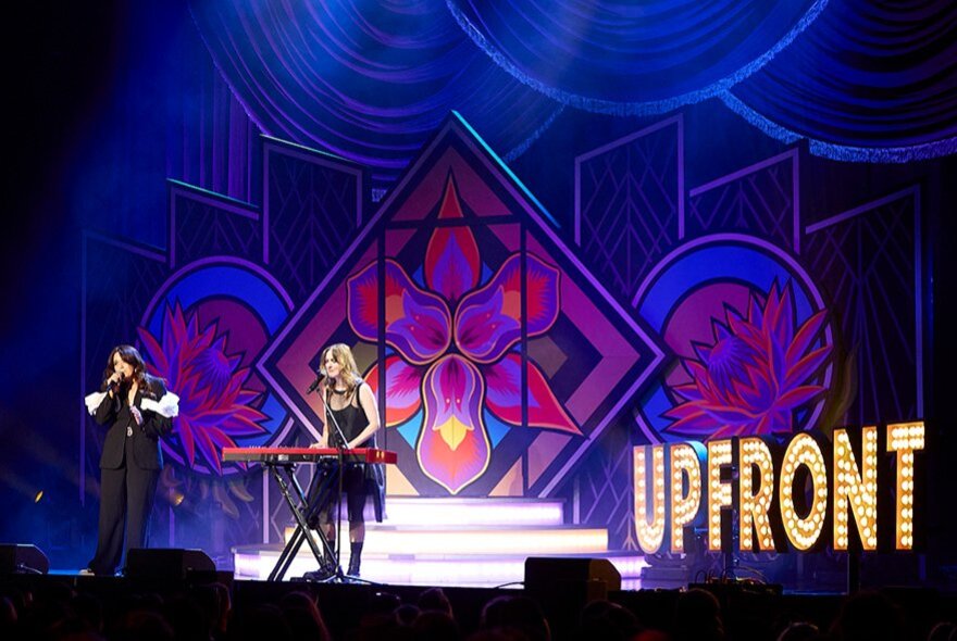 Two performers standing on a stage, one behind a digital keyboard, behind them is a sign spelling out the words UPFRONT in bright lights.