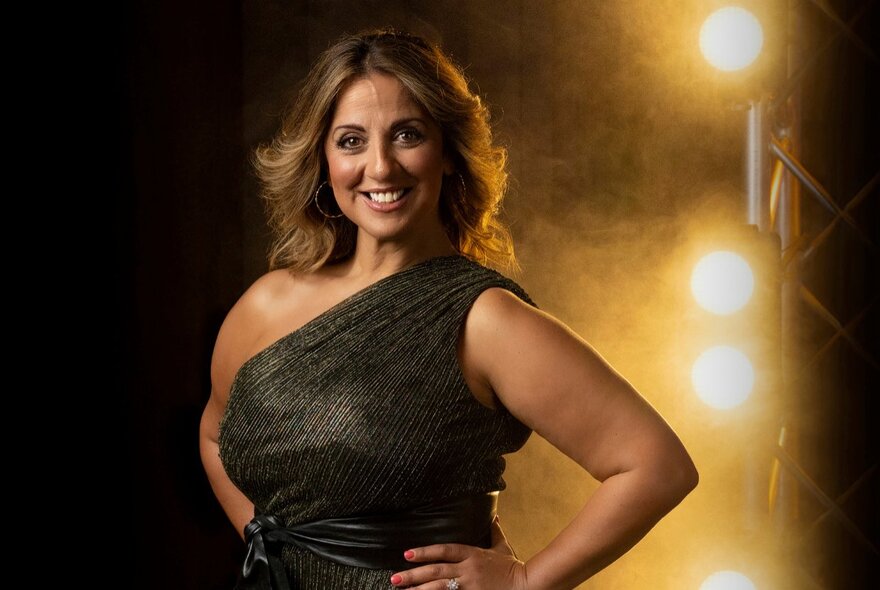 Portrait of Australian musical theatre star Silvie Paladino as she stands with her hands on her hips in a formal gown, in front of bright stage lighting.
