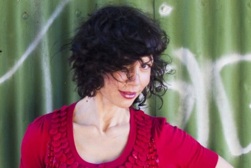 Singer Jackie Bornstein, one eye hidden by her dark hair, wearing a red frilled top, standing in front of green graffitied corrugated iron fence.
