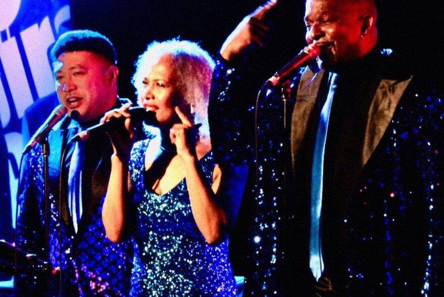 A female and two male singers wearing sparkly outfits performing live on stage.