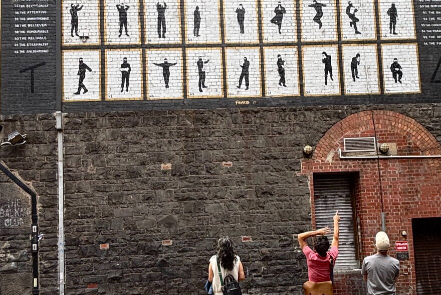 A tour guide wearing a pink t-shirt, pointing to stencil art on the wall above him while talking to a couple on a walking tour.