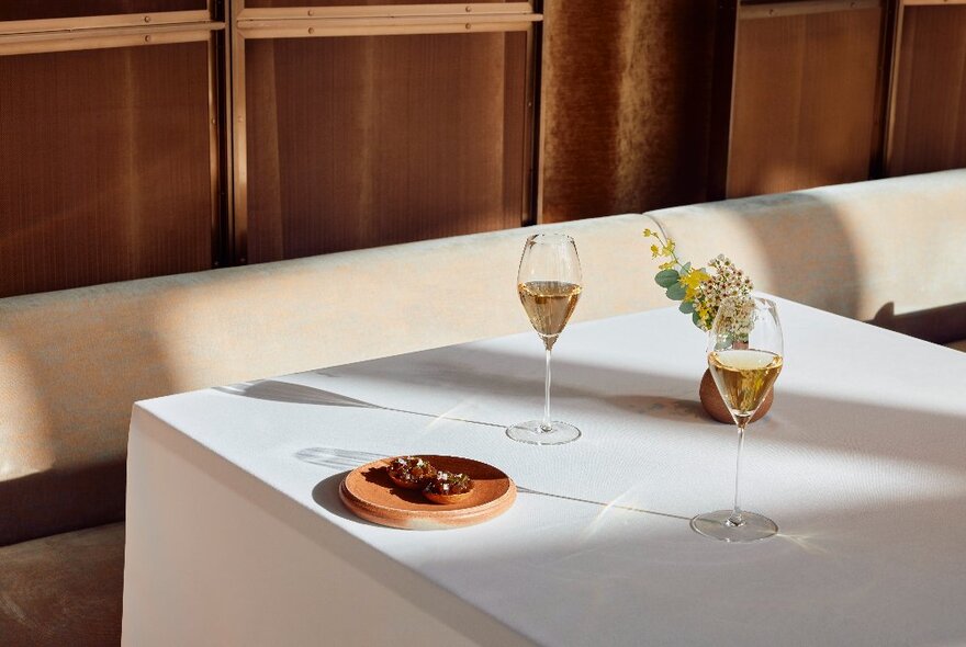 A table with a white tablecloth, two glasses on white wine and a small plate of food.