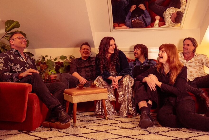 A group of six people, members of a band, seated on sofas and interacting with one another in a room.