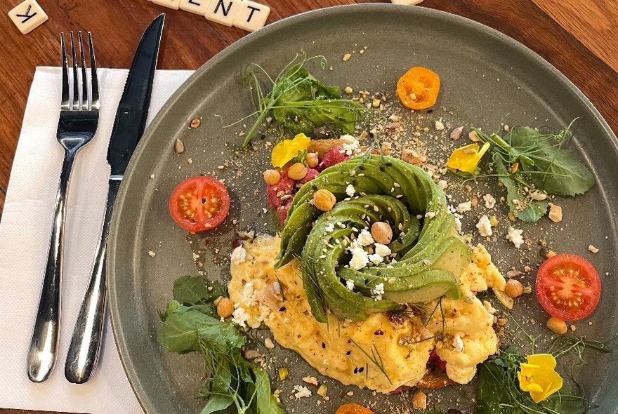 Overhead view of a plate of scrambled eggs with sliced avocado garnish, cherry tomatoes and fresh herbs.