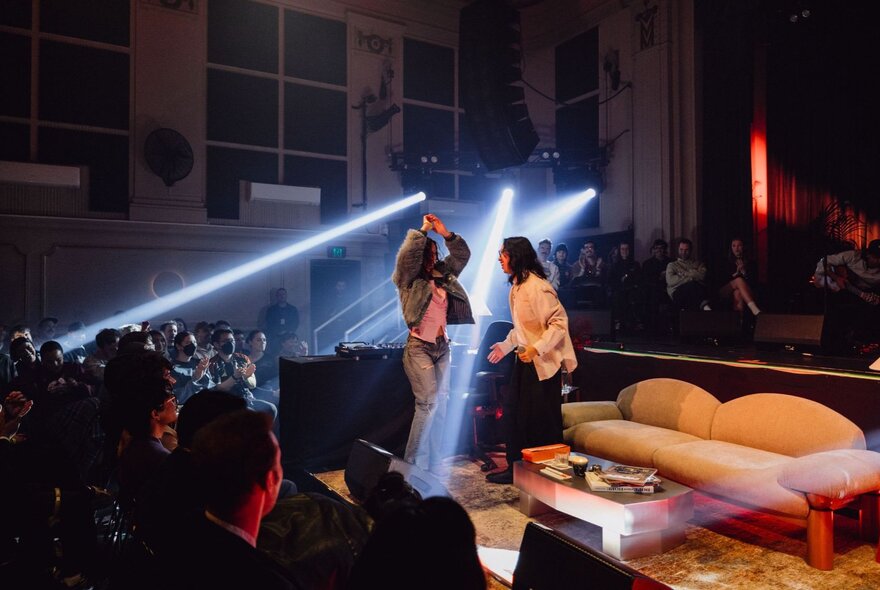 Two people standing near a couch and low table on a stage, the area is spotlit and an audience is watching and listening to them.