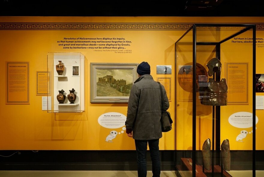 Installation view of the Spartans & Anzacs exhibition at the Shrine of Remembrance, with a person looking at a wall mounted display behind glass.