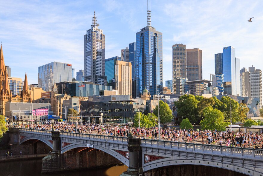 A large group of people taking part in the Melbourne marathon and running across Princess Bridge with tall city buildings and trees in the background, set against a blue sky.