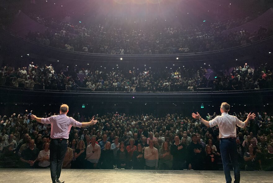 Looking behind two performers on stage with their hands up at the audience. 