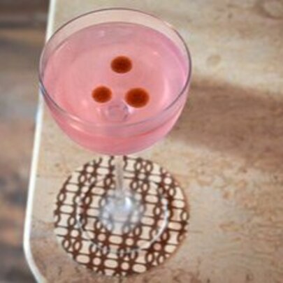 A pink cocktail with three red dots on top, sitting on a checked coaster.