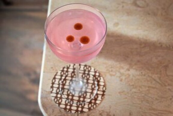 A pink cocktail with three red dots on top, sitting on a checked coaster.