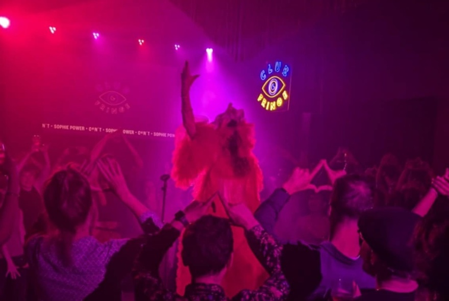 A performer on a stage in front of a audience who are interacting with their arms raised in the air and making signs with their hands.