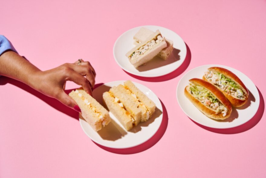 A pink table with three plates of finger sandwiches and a hand reaching for one of the plates. 